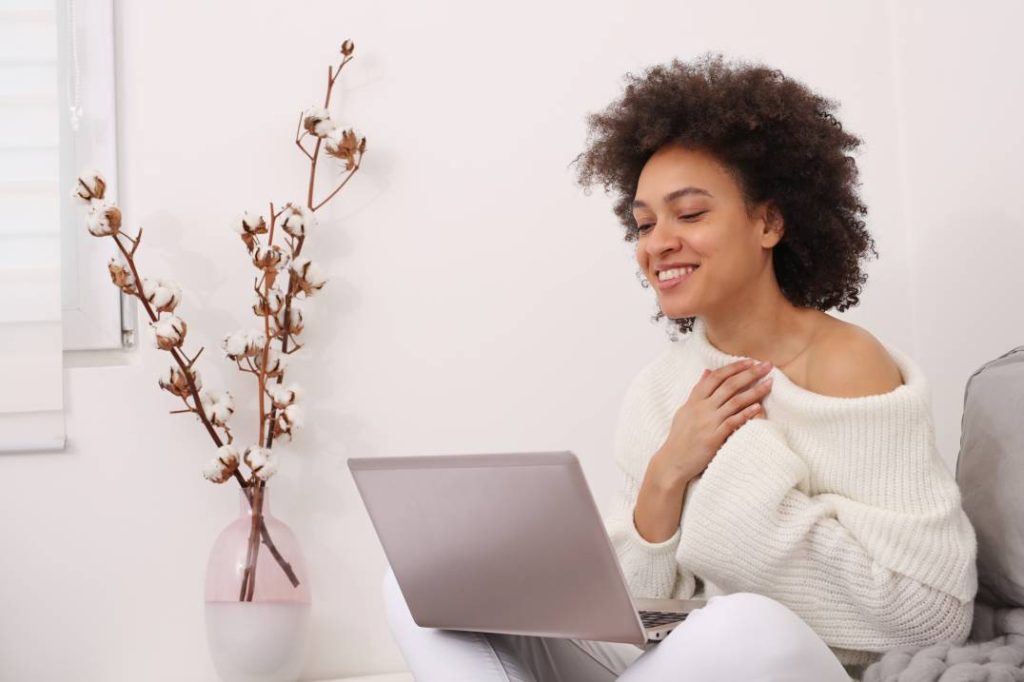 A woman sitting on a couch, focused on her laptop, with a cozy living room setting in the background.